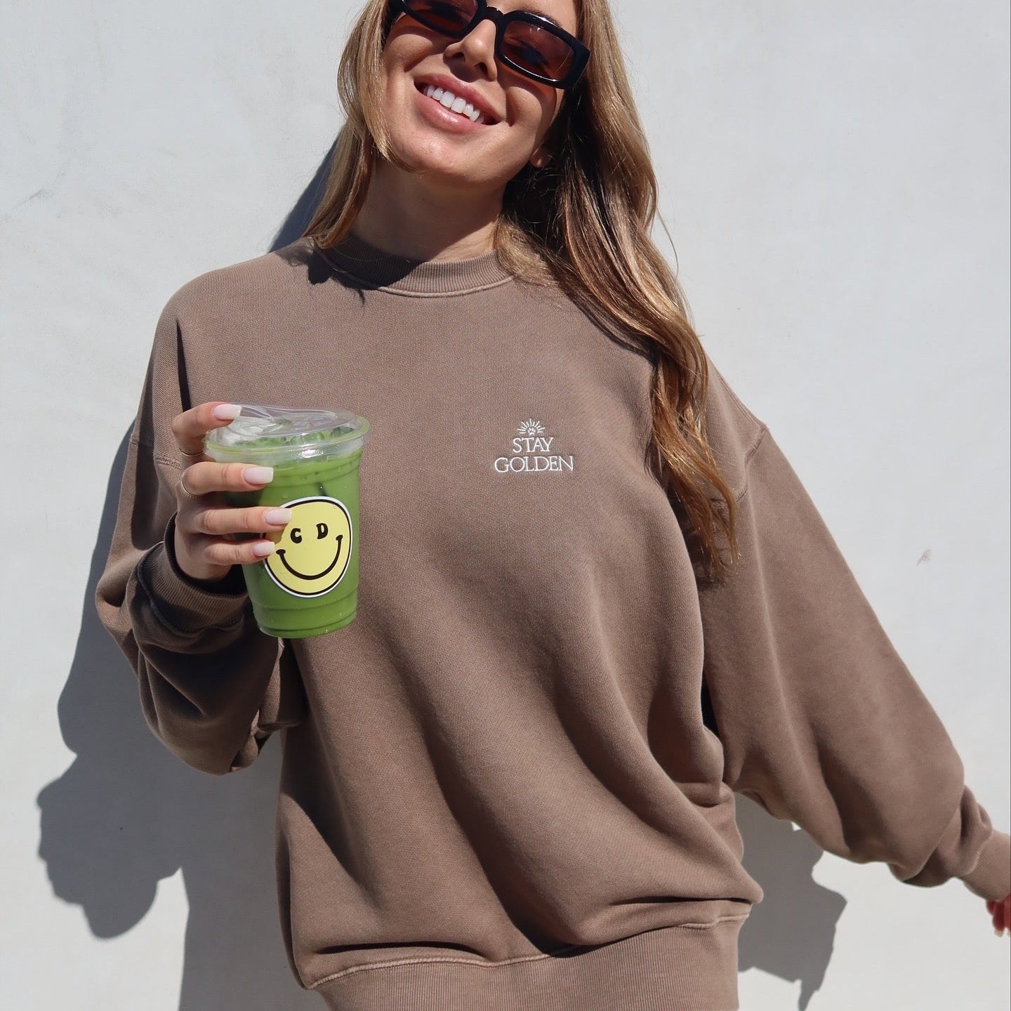 Woman wearing a brown sweatshirt with a logo, holding a green drink with a smiley face, against a light background.