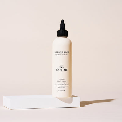 A bottle of Goldie Mercle Rinse hair care product with a black dropper cap, standing on a white rectangular pedestal against a warm beige background.