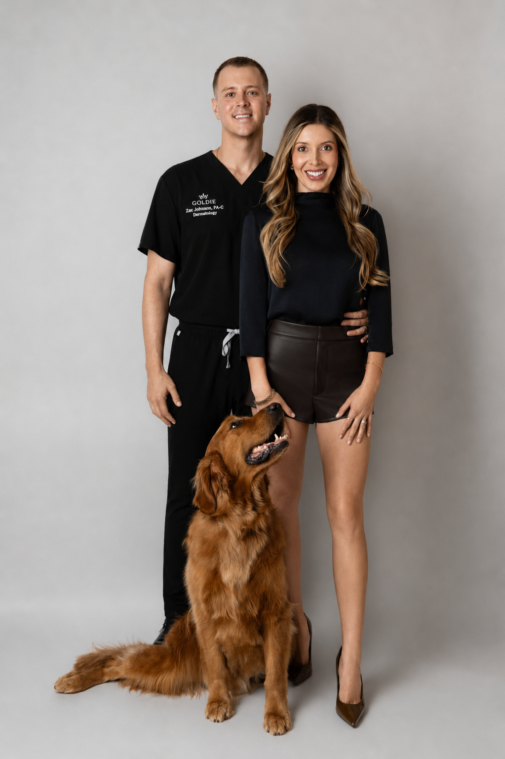 A man in black medical scrubs stands next to a woman in a black top and brown leather skirt, with a golden retriever dog sitting in front of them against a light gray studio background.