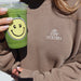 Person wearing a brown sweatshirt with 'Stay Golden' logo, holding a green cup with a smiley face.